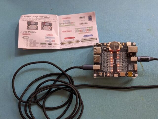 USB Kabeltester mit leuchtenden LEDs, eingestecktem USB-C Kabel und offenem Handbuch.