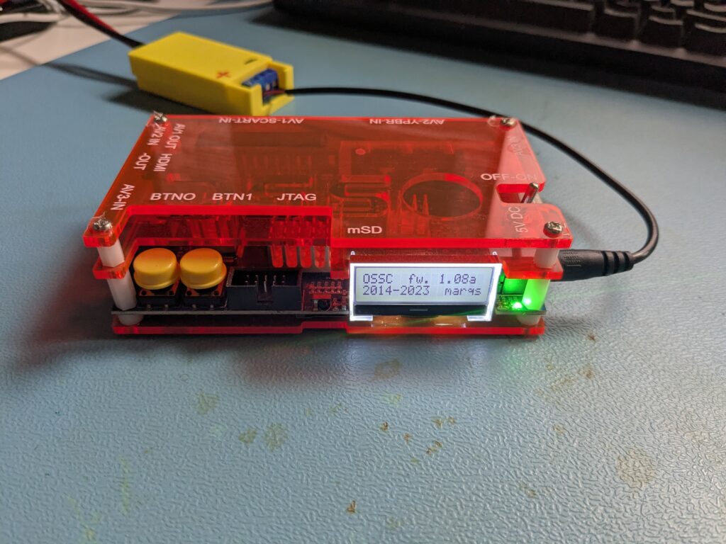 Seitenansicht des OSSC im roten Acrylgehäuse mit gelbem SCART-Adapter oben auf dem Gerät, angeschlossenes Netzteil und LCD-Display mit Firmware 1.08a