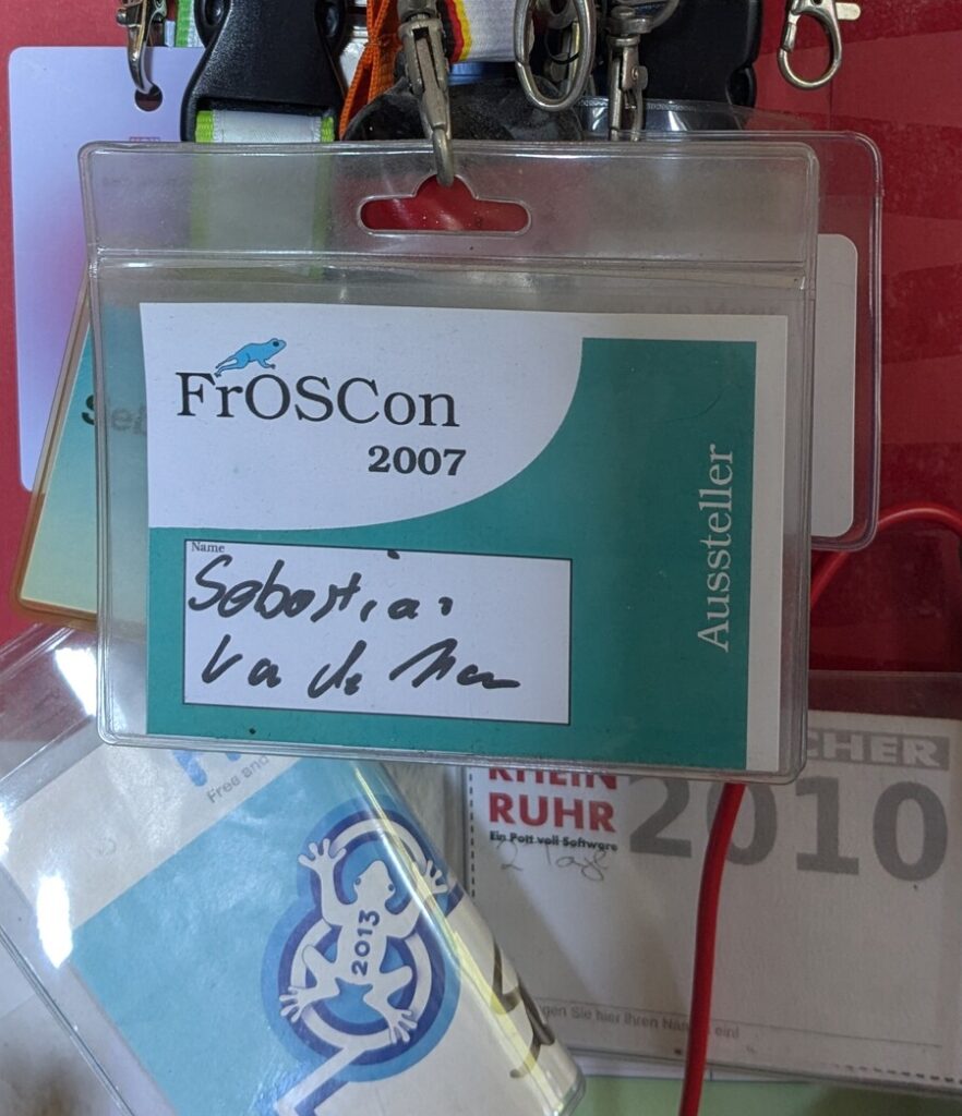 Drei Konferenzausweise an bunten Schlüsselbändern an einer Wand. Vorne der Ausweis der FrOSCon 2007 mit handgeschriebenem Namen und dem senkrechten Aufdruck Aussteller, darunter ein hellblaues Schild der FrOSCon 2013 mit Frosch-Logo, rechts ein Besucherausweis der OpenRheinRuhr 2010.