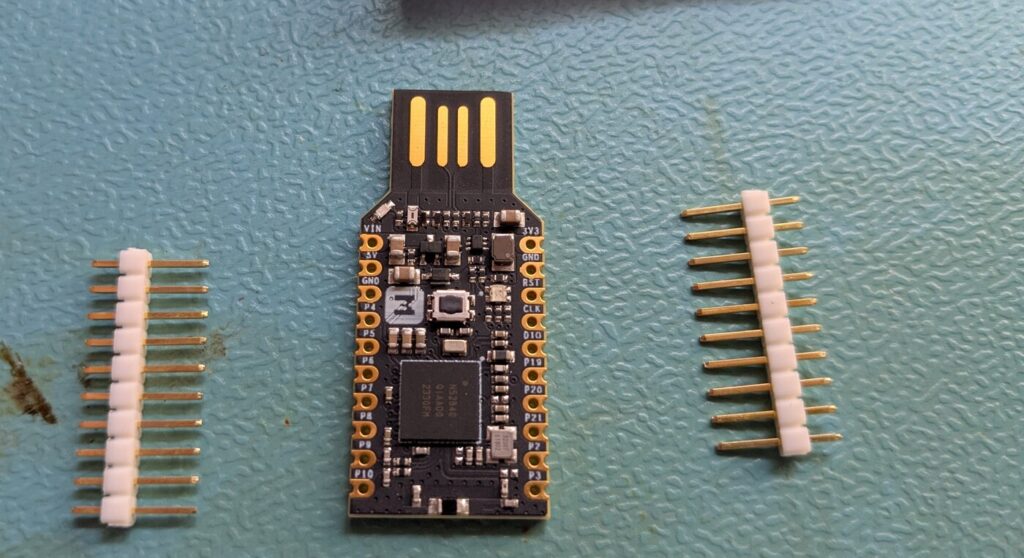 Das makerdiary nRF52840-MDK USB Dongle neben den beiliegenden, losen Pfostenstiftleisten. Auf dem Chip steht der Aufdruck N52840 2330FM.